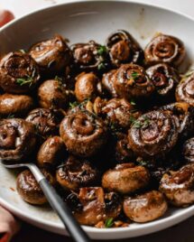 Image shows sauteed steakhouse mushrooms served in a white plate with thyme garnish.