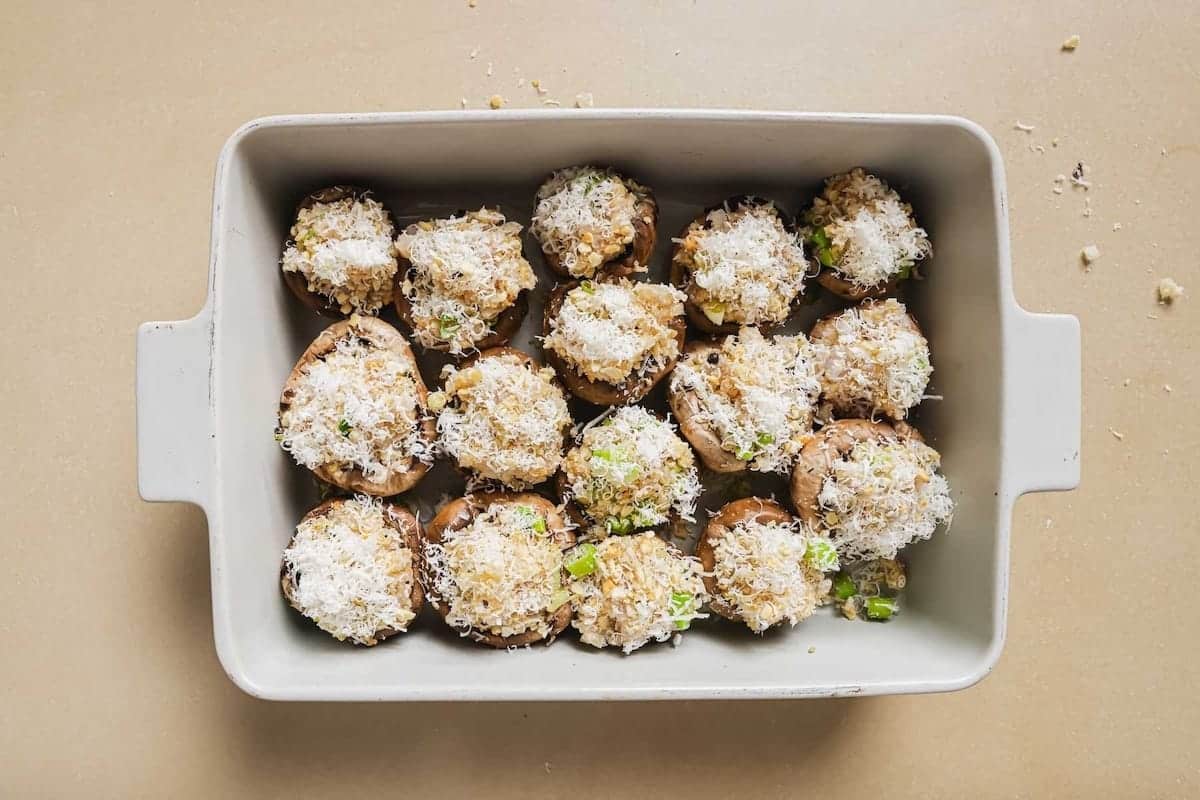 A rectangular baking dish filled with shrimp stuffed mushrooms, topped with grated cheese and green herbs, arranged in a single layer on a light-colored surface.