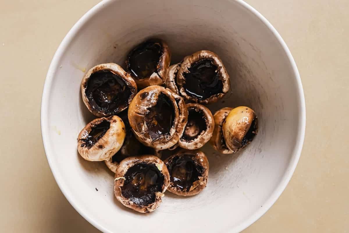 A white bowl containing raw mushroom caps with their stems removed, ready to be transformed into delicious shrimp stuffed mushrooms, sits on a light beige surface.