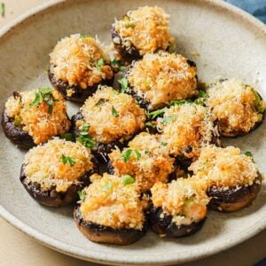 A plate of stuffed mushrooms with shrimp, topped with golden, melted cheese and chopped herbs, arranged neatly on a light-colored dish.