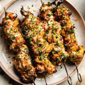 Bang bang chicken skewers topped with a creamy bang bang sauce and chopped herbs, served on a round plate. The skewers are slightly charred and garnished with green chives and red pepper flakes.