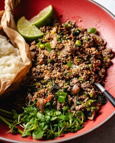 Image shows Thai beef larb cooked to perfectly seasoned with loads of herbs and lime juice. Served on a big red color plate with sticky rice.