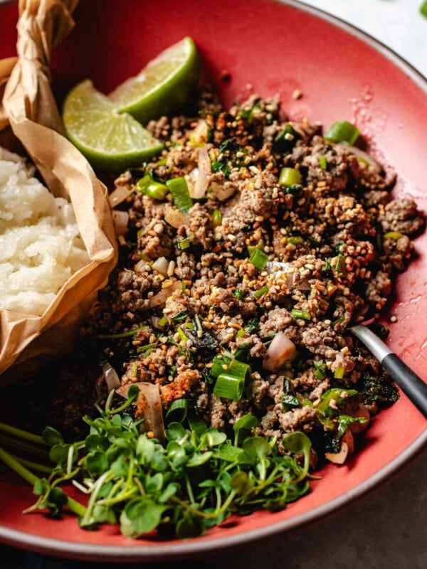 Image shows Thai beef larb cooked to perfectly seasoned with loads of herbs and lime juice. Served on a big red color plate with sticky rice.