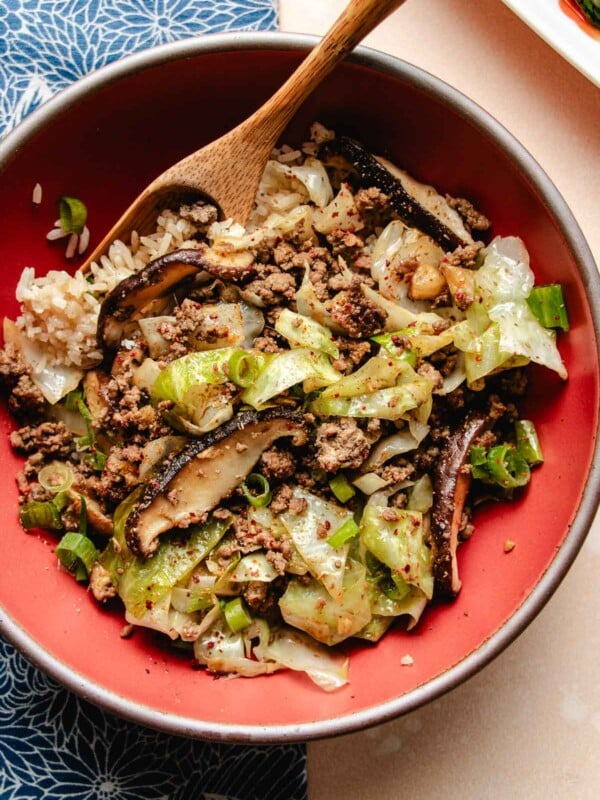 A close overhead shot shows ground beef stir fried to perfectly browned with cabbage and shiitake and served with rice in a red bowl.