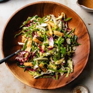 Recipe image shows napa cabbage slaw with other thinly shredded vegetables and crunchy toppings served in a wood plate with dressing on top.