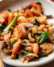 A close shot shows juicy shrimp stir fried with cashew, ginger, garlic, scallions with shacha sauce and served on a white plate.