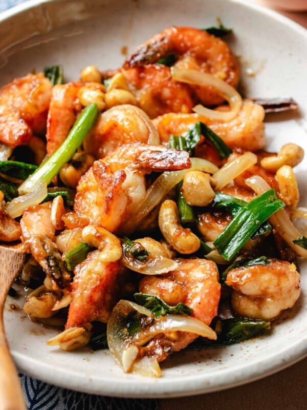 A close shot shows juicy shrimp stir fried with cashew, ginger, garlic, scallions with shacha sauce and served on a white plate.