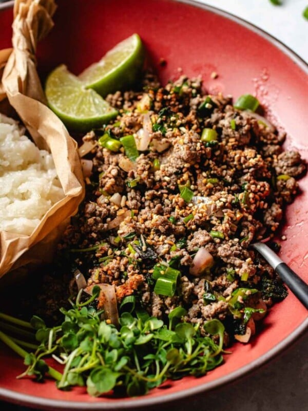 Image shows Thai beef larb cooked to perfectly seasoned with loads of herbs and lime juice. Served on a big red color plate with sticky rice.