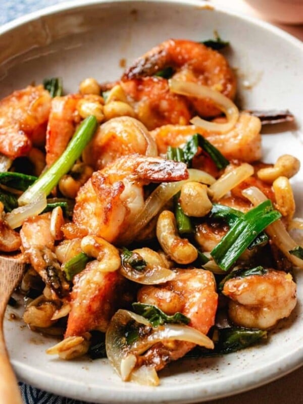 A close shot shows juicy shrimp stir fried with cashew, ginger, garlic, scallions with shacha sauce and served on a white plate.