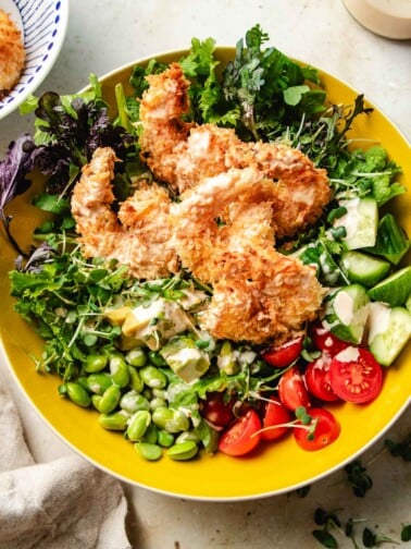 An overhead image shows crunchy bang bang shrimp served with a bowl of colorful fresh greens and vegetables in a yellow bowl.
