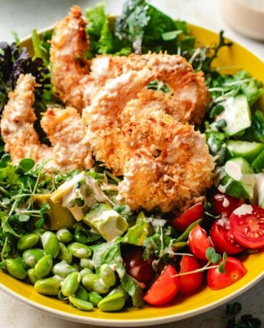 Image shows a side close shot with crispy bang bang shrimp on top of a bowl of salad greens and vegetables, served in a yellow color bowl.