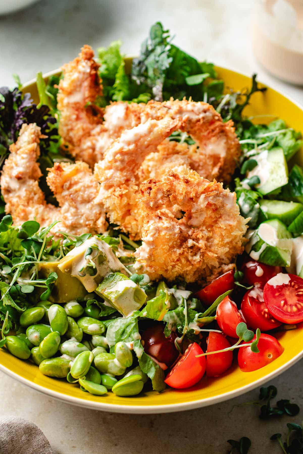 Image shows a side close shot with crispy bang bang shrimp on top of a bowl of salad greens and vegetables, served in a yellow color bowl.