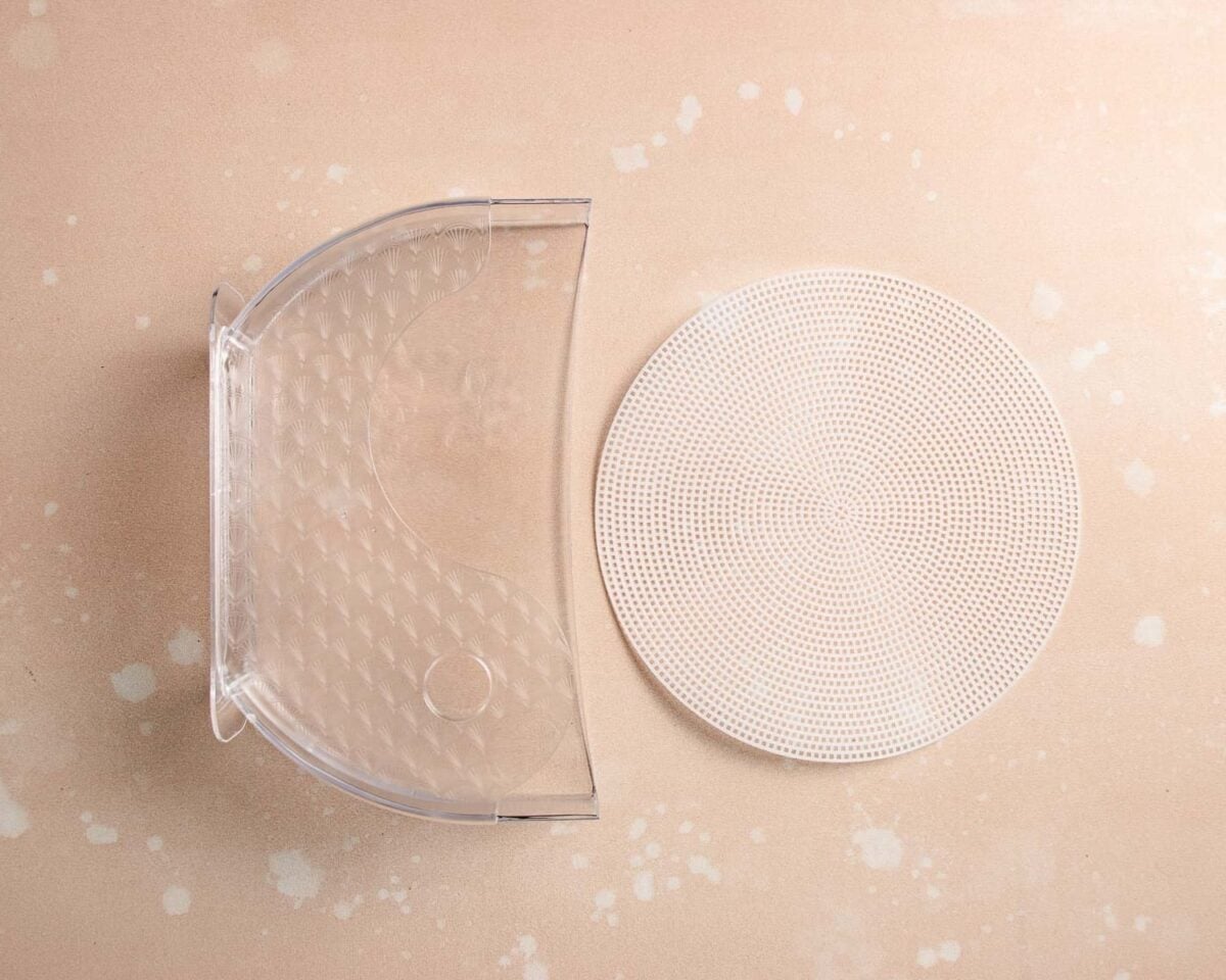 A clear plastic rice paper water dipping bowl sits next to a round, flat, white perforated mat—ideal for use with a rice paper wrapping tray—on a beige speckled surface.