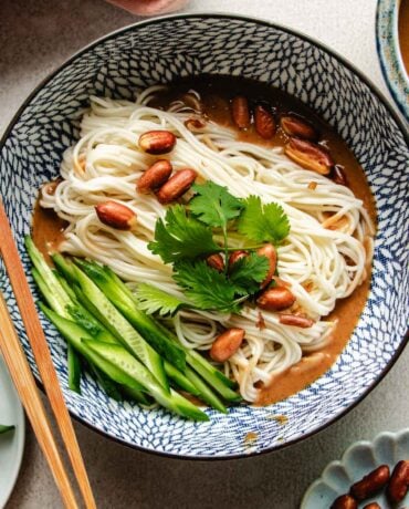 An overhead shot image shows a blue and white color bowl with boiled cold noodles inside with Peanut butter sauce and veggies on the side.