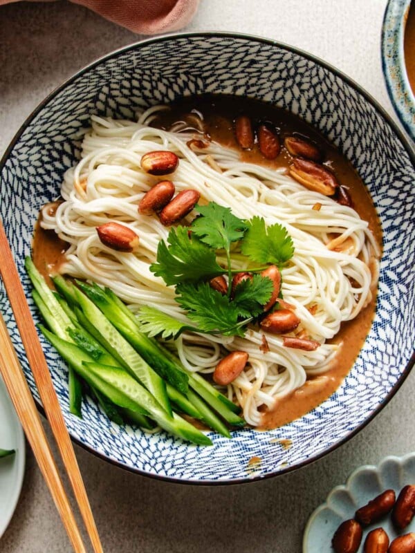 An overhead shot image shows a blue and white color bowl with boiled cold noodles inside with Peanut butter sauce and veggies on the side.