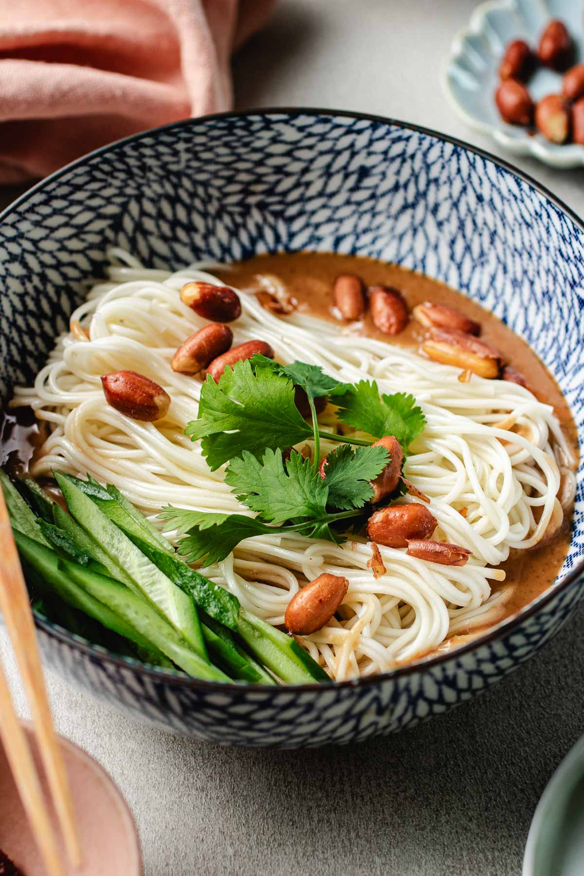A side image shows beautiful boiled and chilled cold noodles served in an Asian blue white color noodle bowl with peanut sauce at the bottom and veggies on the side.