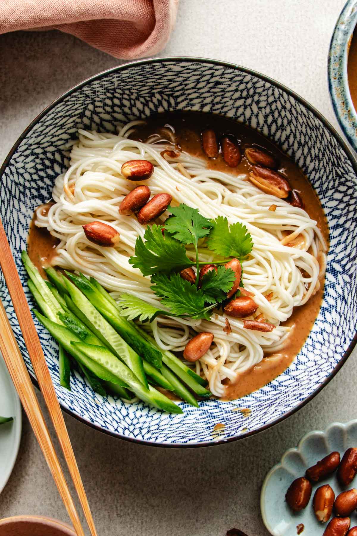 An overhead shot image shows a blue and white color bowl with boiled cold noodles inside with Peanut butter sauce and veggies on the side.