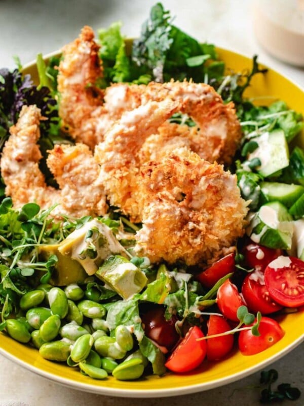 Image shows a side close shot with crispy bang bang shrimp on top of a bowl of salad greens and vegetables, served in a yellow color bowl.