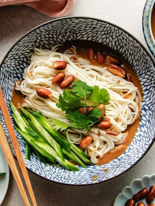 An overhead shot image shows a blue and white color bowl with boiled cold noodles inside with Peanut butter sauce and veggies on the side.