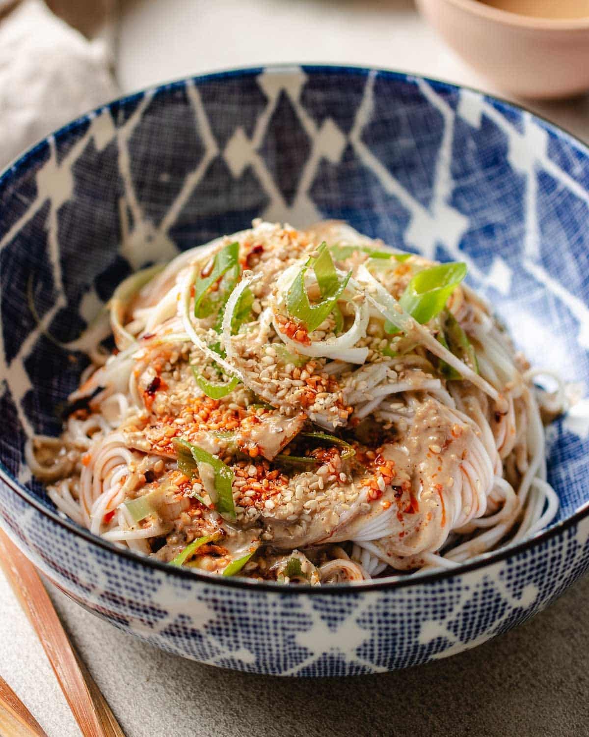 Feature image shows a bowl of cold noodles covered in Taiwanese sesame sauce served in a blue white color bowl with sesame seeds on top.