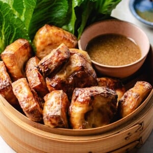 Feature image shows tofu puffs with golden brown color outside and fluffy inside, served in a bamboo basket with dipping sauce and lettuce greens on the side.