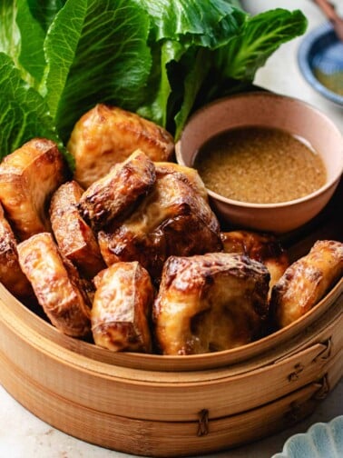 Feature image shows tofu puffs with golden brown color outside and fluffy inside, served in a bamboo basket with dipping sauce and lettuce greens on the side.