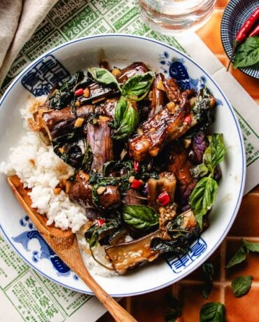 Image shows Thai basil eggplant coated with sweet basil sauce, served with rice in a blue white color bowl.