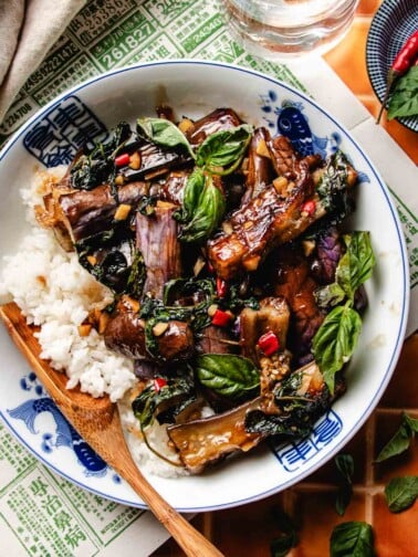 Image shows Thai basil eggplant coated with sweet basil sauce, served with rice in a blue white color bowl.