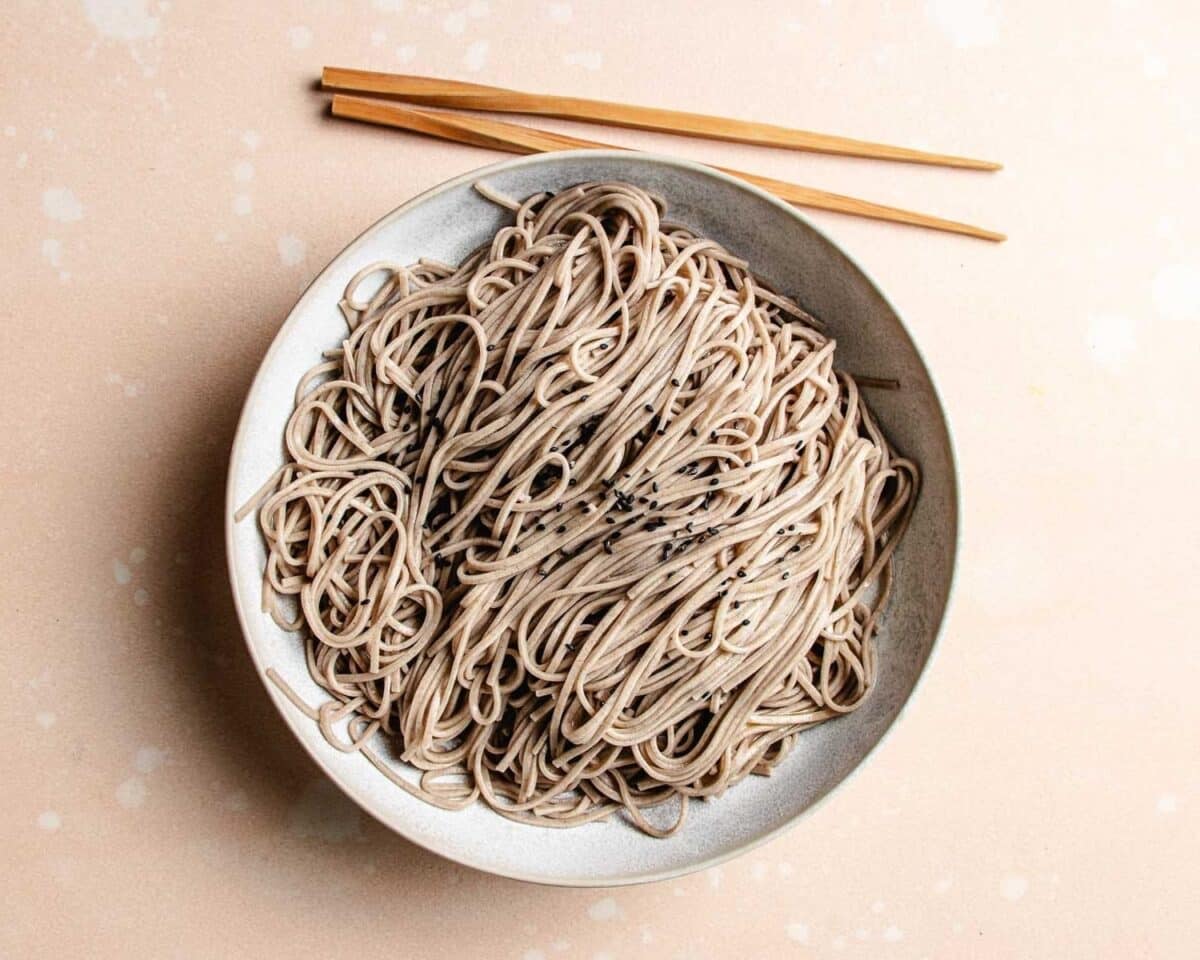 A bowl of plain soba noodles topped with black sesame seeds, perfect for an Asian noodle salad, placed on a light surface with a pair of wooden chopsticks resting above the bowl.