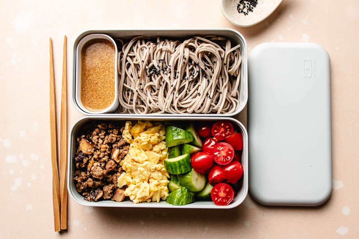 A bento box with compartments holds an Asian noodle salad featuring soba noodles topped with sesame seeds, a small container of sauce, chopped cucumbers, cherry tomatoes, scrambled eggs, and ground meat with mushrooms. Wooden chopsticks lie beside the box.