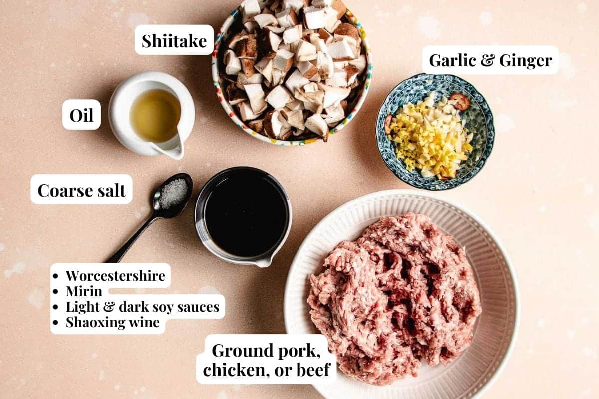 Ingredients for a savory soba noodle salad recipe are arranged on a light surface: chopped shiitake mushrooms, minced garlic and ginger, oil, coarse salt, sauces, seasonings, and a bowl of ground pork, chicken, or beef. Labels identify each item.