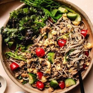 A vibrant soba noodle salad, this cold soba noodle salad recipe features soba noodles tossed with ground meat, scrambled eggs, cherry tomatoes, cucumber, and leafy greens, topped with black sesame seeds. A black fork rests in the bowl.