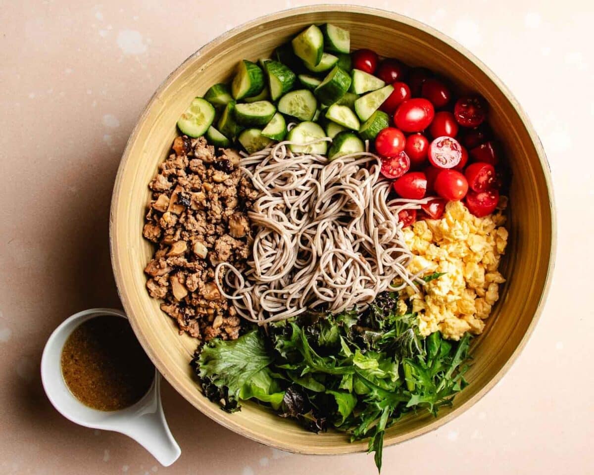 A large wooden bowl filled with chopped cucumber, cherry tomatoes, scrambled eggs, cooked ground meat, soba noodles, and mixed greens creates a vibrant Asian noodle salad. A small pitcher of dressing sits beside the bowl.