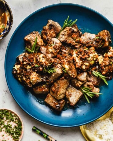 Feature image shows tender garlic steak bites top with loads of garlic and garnished with rosemary, served on a blue color serving plate.