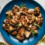 Recipe image shows garlic steak bites served on a big blue color plate and garnished with garlic and rosemary on top.