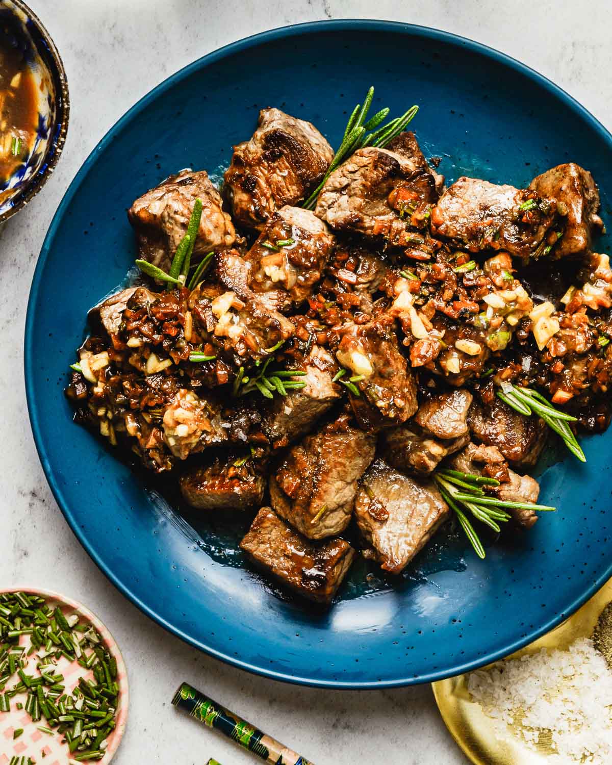 Feature image shows tender garlic steak bites top with loads of garlic and garnished with rosemary, served on a blue color serving plate.