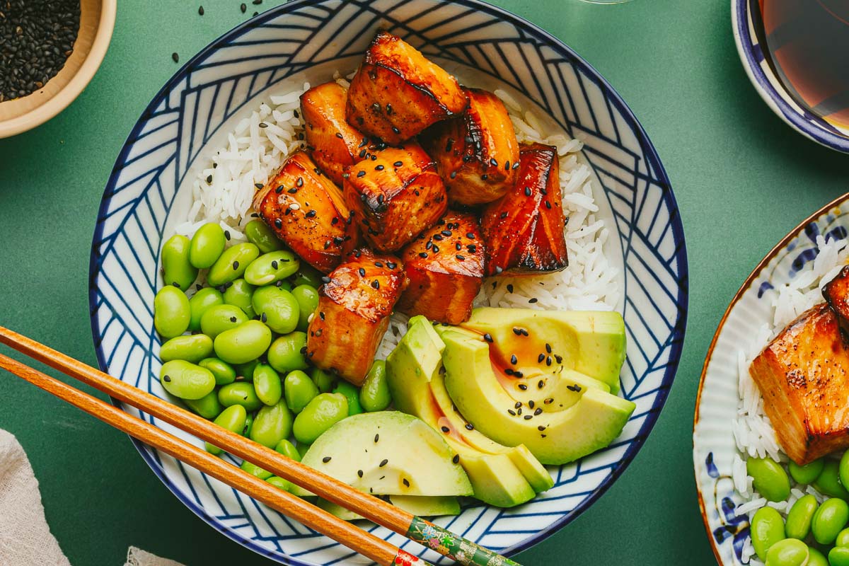 A horizontal image shows two bowls of teriyaki salmon bites served with rice, edamame, and avocado and gluten free teriyaki sauce on the side.