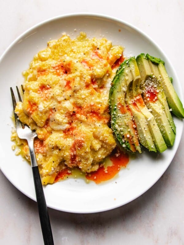 Feature image shows a plate of creamy scrambled eggs made in an air fryer and serve with avocado and hot sauce on top.