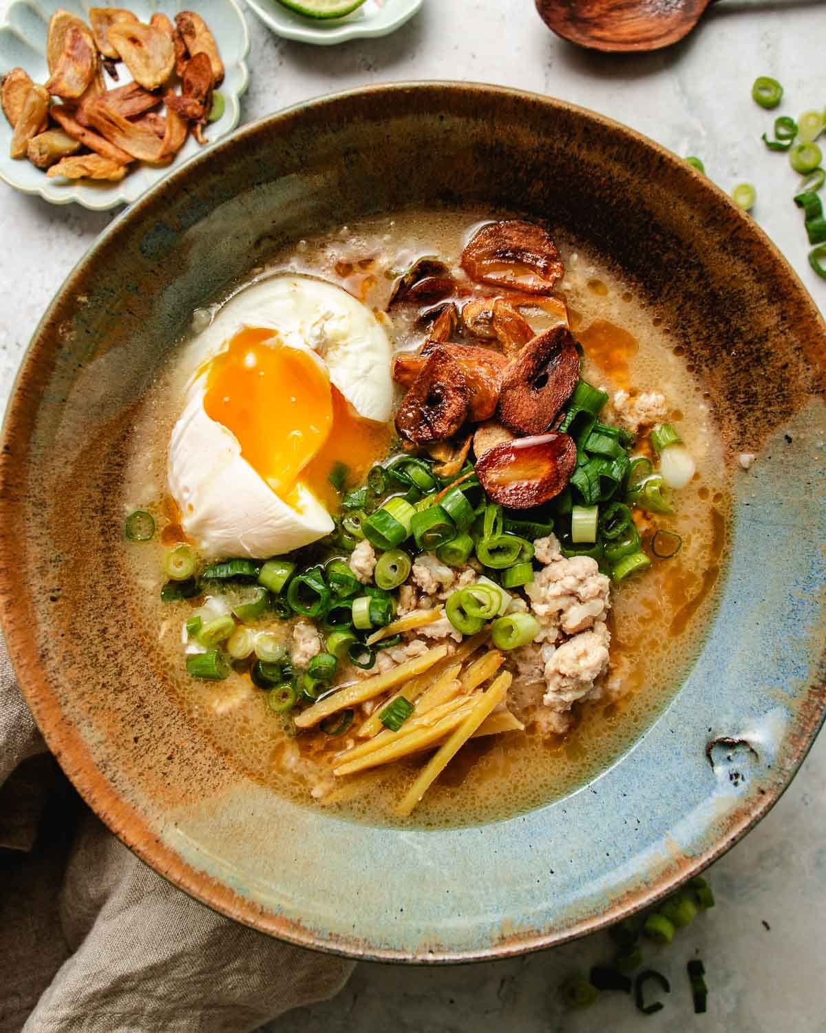 An overhead feature image shows a bowl of delicious Creamy chicken and rice soup Taiwanese style with garlic chips, ginger, green onions, and a poached egg served in one bowl.