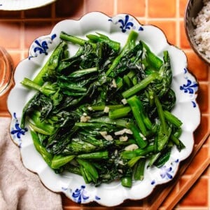 Photo shows a plate of stir fried choy sum with garlic, served over a blue white color plate with chopsticks on the side.
