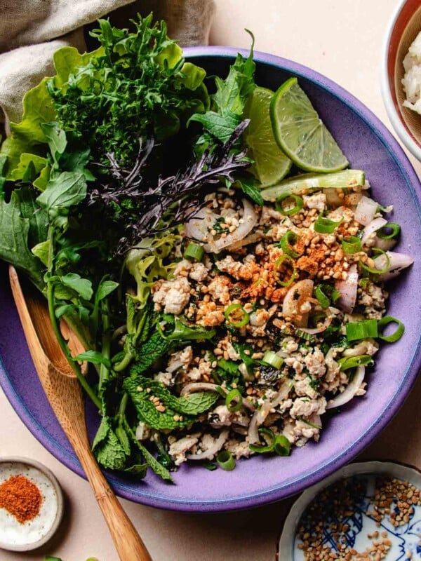 Feature image shows Thai chicken larb with salad greens on the side and topped with toasted rice powder and chili powder, served over a purple color plate.