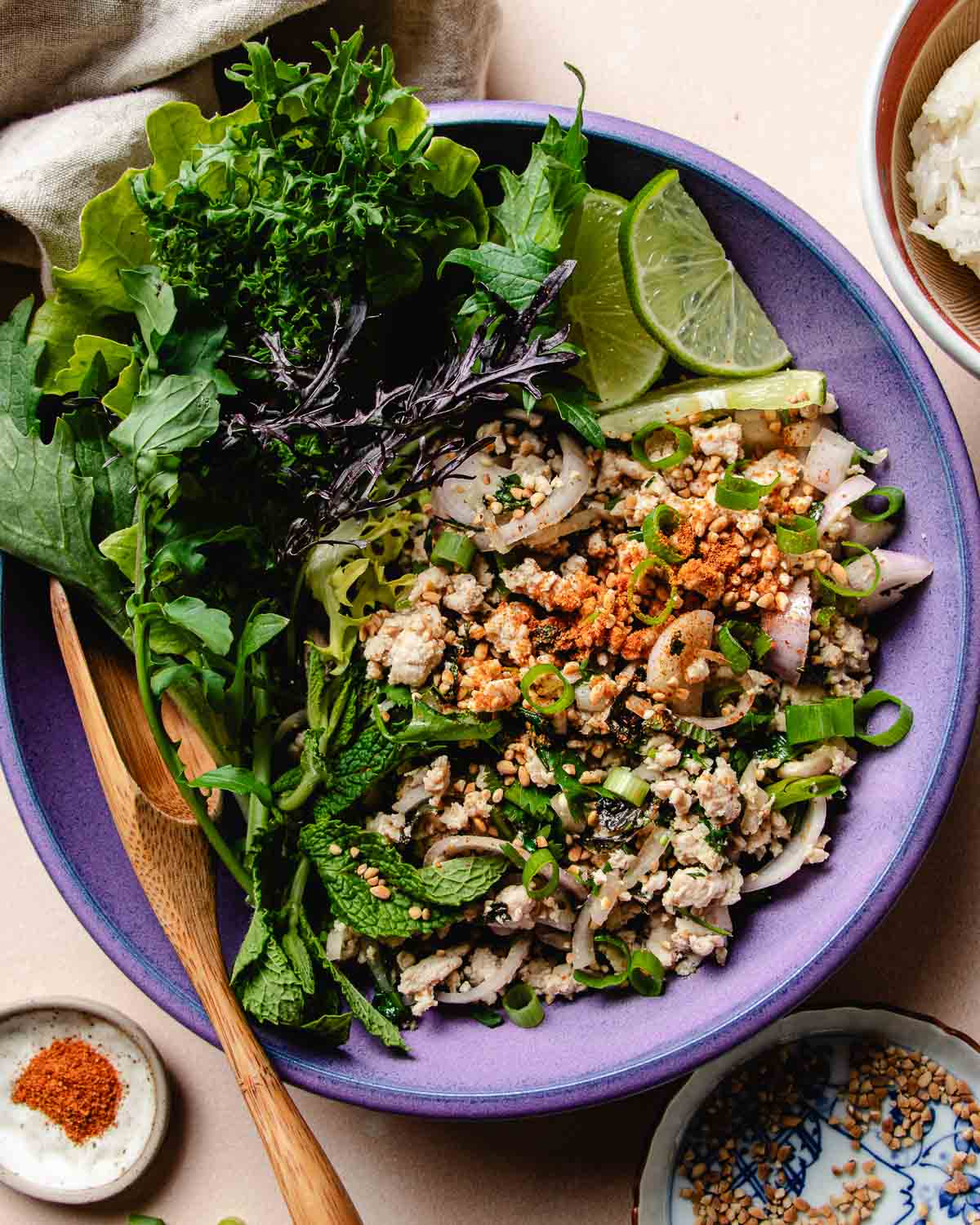 Feature image shows Thai chicken larb with salad greens on the side and topped with toasted rice powder and chili powder, served over a purple color plate.