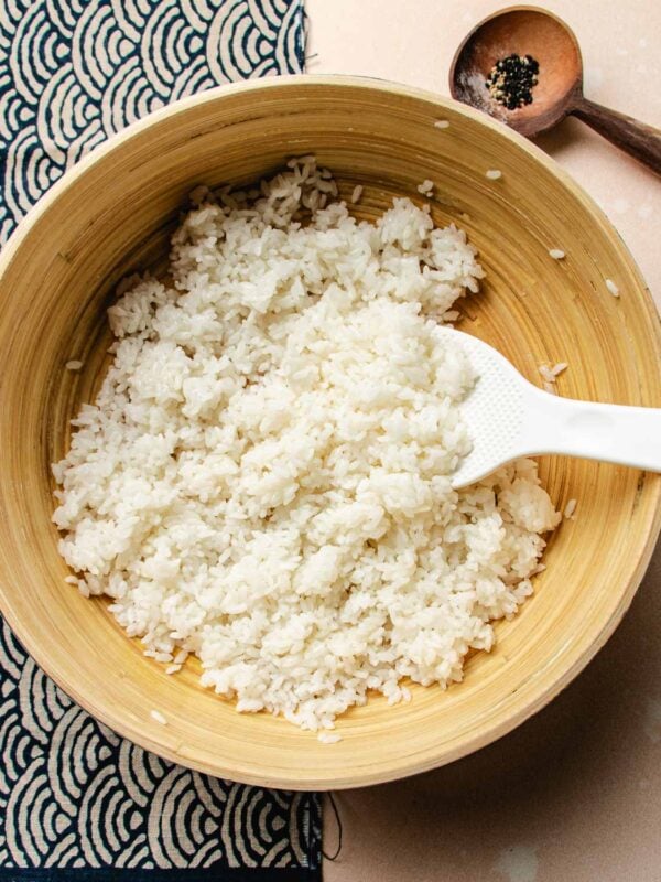 Overhead feature image shows freshly cooked sushi rice seasoned with sushi vinegar in a big bamboo mixing bowl with rice paddle on the side.