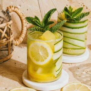 Two glasses of a refreshing yellow-green cucumber mocktail, garnished with mint, lemon slices, and cucumber ribbons, sit on white coasters as sunlight casts warm shadows across a beige stone surface.