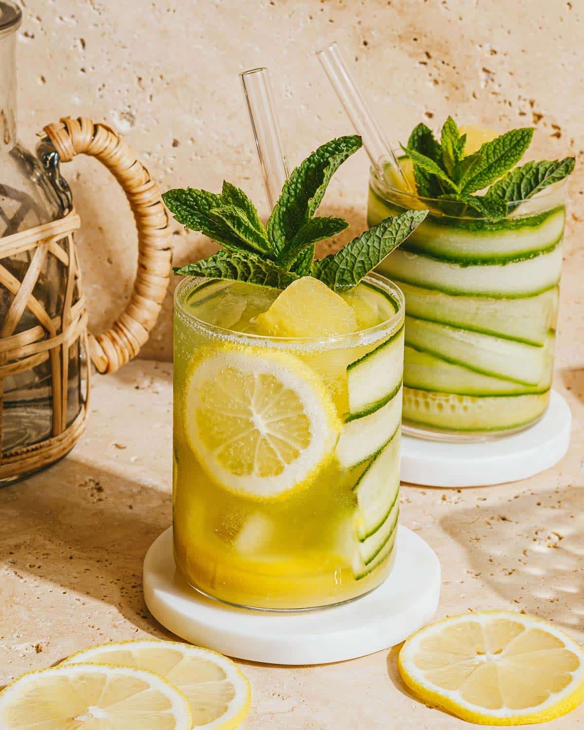 Two refreshing cucumber mocktails garnished with lemon slices, cucumber ribbons, and fresh mint leaves, served in clear glasses with straws on white coasters. Lemon slices and a pitcher are nearby on a beige surface.