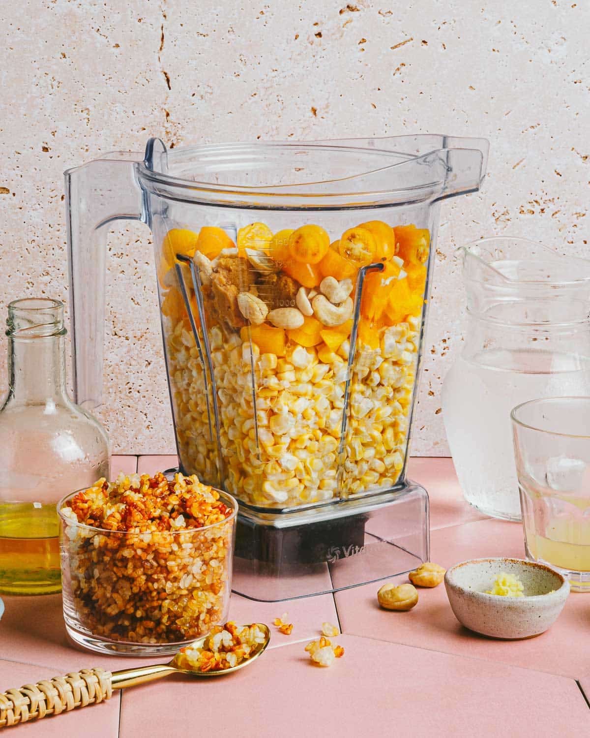 A blender filled with corn, yellow cherry tomatoes, cashews, and other ingredients for Corn Gazpacho stands on a pink countertop, surrounded by jars of oil, water, a glass, a bowl of seasoning, and a spoon with crushed spices.