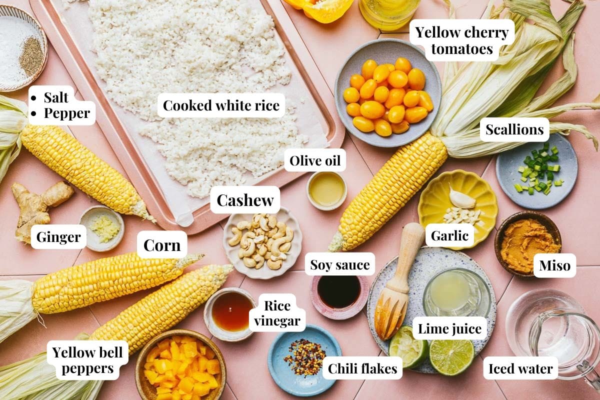 A flat lay of Corn Gazpacho ingredients on a pink surface, including cooked white rice, corn, yellow cherry tomatoes, peppers, scallions, ginger, cashews, miso, soy sauce, lime juice, and more. Each item is clearly labeled.