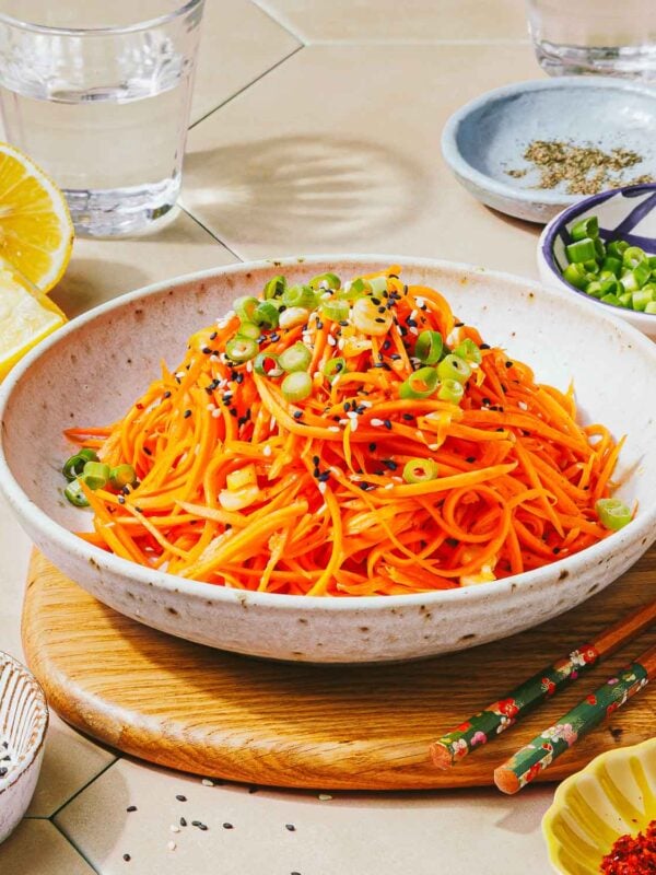 A bowl of Korean carrot salad, topped with sliced green onions and black and white sesame seeds, sits on a wooden board. Lemon wedges, chopped green onions, spices, and chopsticks are placed nearby.