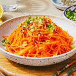 A bowl of Korean carrot salad topped with sliced green onions and black sesame seeds, placed on a wooden surface with chopsticks and small dishes of condiments nearby.