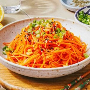 A bowl of Korean carrot salad topped with sliced green onions and black sesame seeds, placed on a wooden surface with chopsticks and small dishes of condiments nearby.